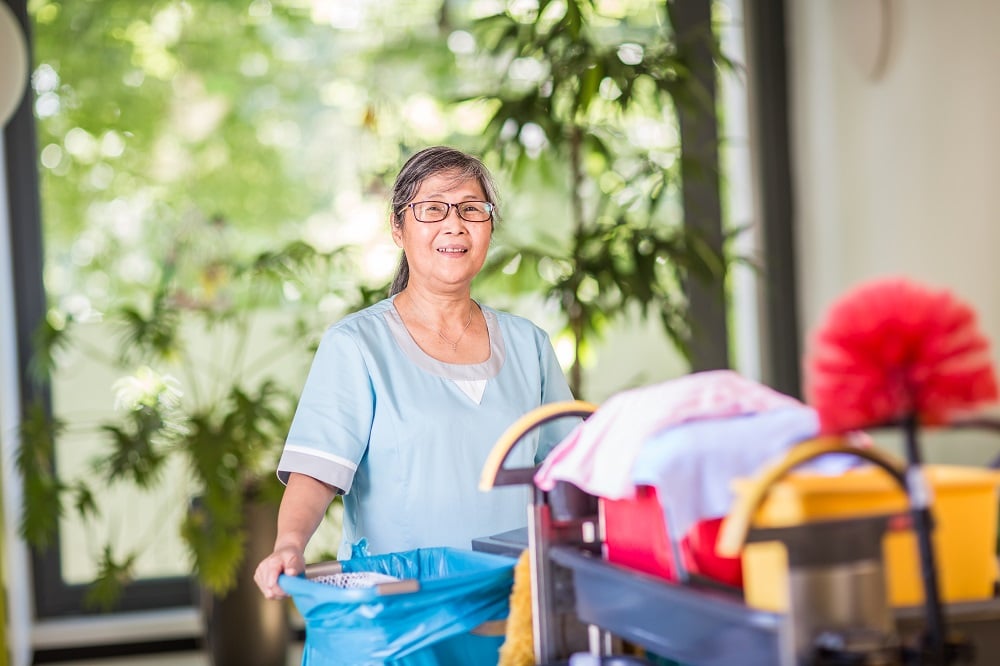 KI-basierte Bildbeschreibung | Eine Frau in Reinigungskleidung steht in einem Innenraum und hält einen blauen Müllsack neben einem Reinigungswagen mit verschiedenen Materialien, mit Pflanzen und großen Fenstern im Hintergrund.